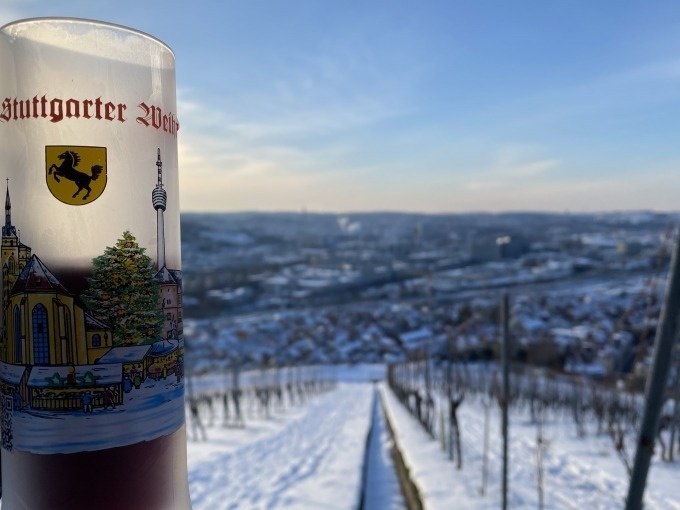Ein Glas Glühwein mit Stuttgarter Motiv steht vor einer verschneiten Weinberglandschaft. Im Hintergrund ist die Stadt Stuttgart zu sehen., © Weingenuss Stuttgart