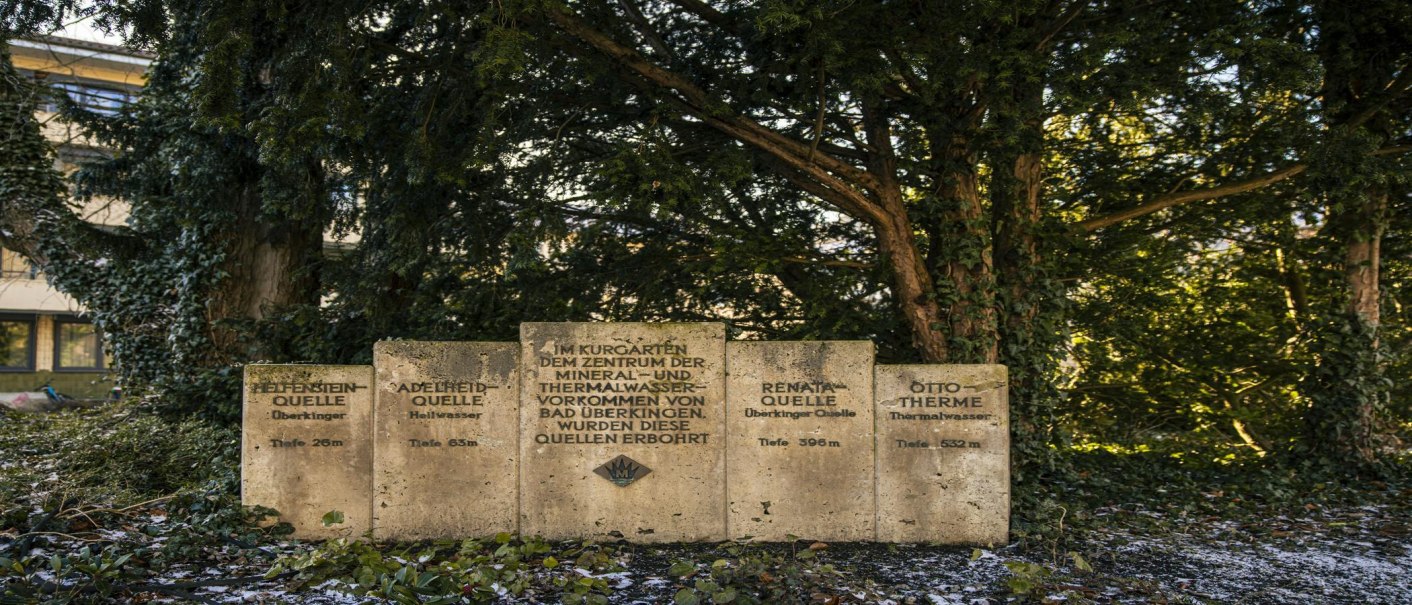 Steintafeln im Kurpark von Bad Überkingen informieren über verschiedene Quellen, umgeben von Bäumen und Efeu., © SMG, Sarah Schmid
