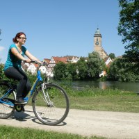 Eine Frau fährt Fahrrad am Neckar in Nürtingen. Im Hintergrund sind eine Kirche und Fachwerkhäuser zu sehen., © Stadt Nürtingen