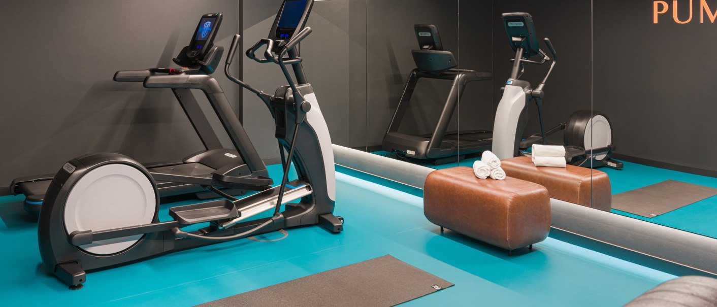 Modernes Fitnessstudio mit Laufband, Crosstrainer, Spiegelwand und blauer Bodenmatte. Ein brauner Hocker mit Handt&uuml;chern steht bereit., &copy; EmiLu Hotel GmbH