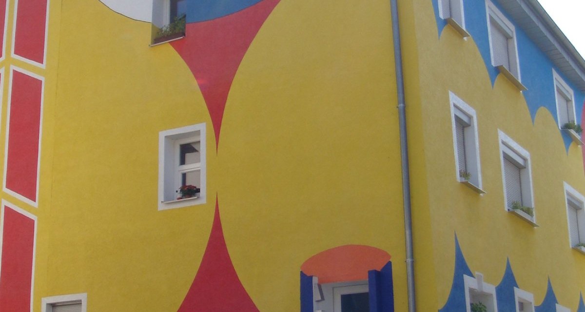 Ein Gebäude mit farbenfroher Fassade, bemalt in Rot, Gelb und Blau, zeigt geometrische Muster und ein großes Auge. Pflanzen schmücken den Eingangsbereich., © Land der 1000 Hügel - Kraichgau-Stromberg Ein Gebäude mit farbenfroher Fassade, bemalt in Rot, Gelb und Blau, zeigt geometrische Muster und ein großes Auge. Pflanzen schmücken den Eingangsbereich., © Land der 1000 Hügel - Kraichgau-Stromberg