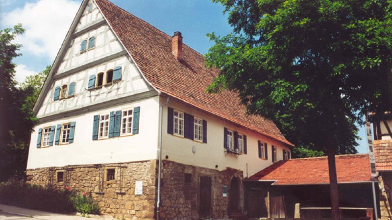 Fachwerkhaus mit rotem Dach und blauen Fensterl&auml;den, umgeben von B&auml;umen. Das Geb&auml;ude steht auf einem steinernen Fundament., &copy; Alfred R&ouml;sner