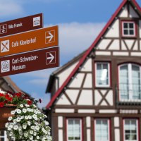 Ein Wegweiser mit Blumen vor Fachwerkhäusern auf dem Marktplatz von Murrhardt. Schilder zeigen zu Villa Franck, Café Klosterhof und Carl-Schweizer-Museum., © Stuttgart-Marketing GmbH, Achim Mende Ein Wegweiser mit Blumen vor Fachwerkhäusern auf dem Marktplatz von Murrhardt. Schilder zeigen zu Villa Franck, Café Klosterhof und Carl-Schweizer-Museum., © Stuttgart-Marketing GmbH, Achim Mende