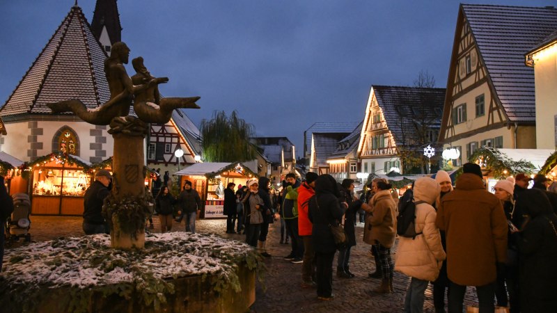 Menschen genießen den Plochinger Weihnachtsmarkt mit beleuchteten Fachwerkhäusern und einer Statue im Vordergrund. Es liegt Schnee auf den Dächern., © Matthias Drißner Menschen genießen den Plochinger Weihnachtsmarkt mit beleuchteten Fachwerkhäusern und einer Statue im Vordergrund. Es liegt Schnee auf den Dächern., © Matthias Drißner