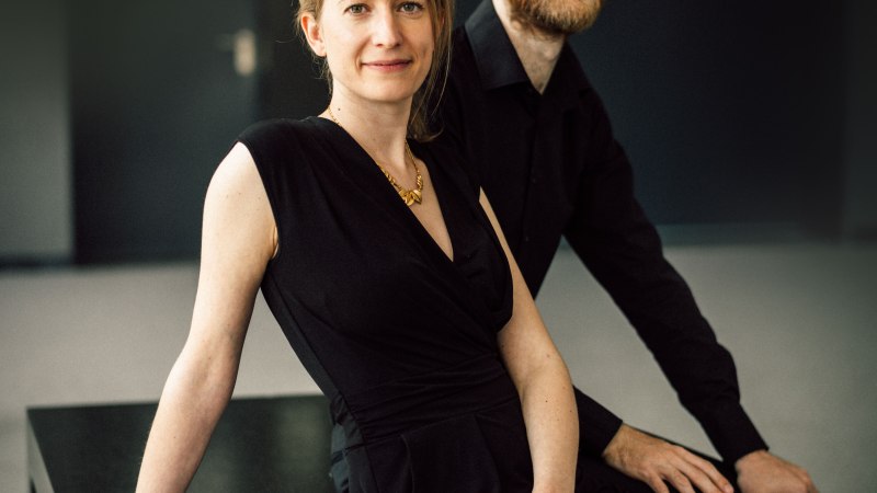 Zwei Personen in eleganter schwarzer Kleidung sitzen auf einem Tisch vor einem dunklen Hintergrund. Sie wirken entspannt und selbstbewusst., © ONYX Klavierduo Zwei Personen in eleganter schwarzer Kleidung sitzen auf einem Tisch vor einem dunklen Hintergrund. Sie wirken entspannt und selbstbewusst., © ONYX Klavierduo