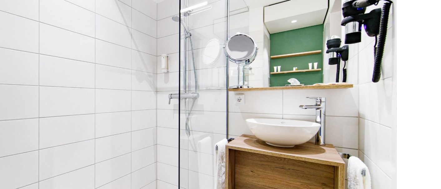 Modernes Badezimmer mit Glasdusche, rundem Waschbecken auf Holzablage, Spiegel und Haartrockner an der Wand. Weiße Fliesen und grüner Akzent an der Wand., © Arthotel ANA Momentum