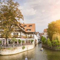 Fachwerkhaus am Roßneckarkanal, umgeben von Bäumen. Menschen sitzen im Café, während Kanufahrer auf dem Wasser unterwegs sind. Sonnenschein im Hintergrund., © Stuttgart-Marketing GmbH