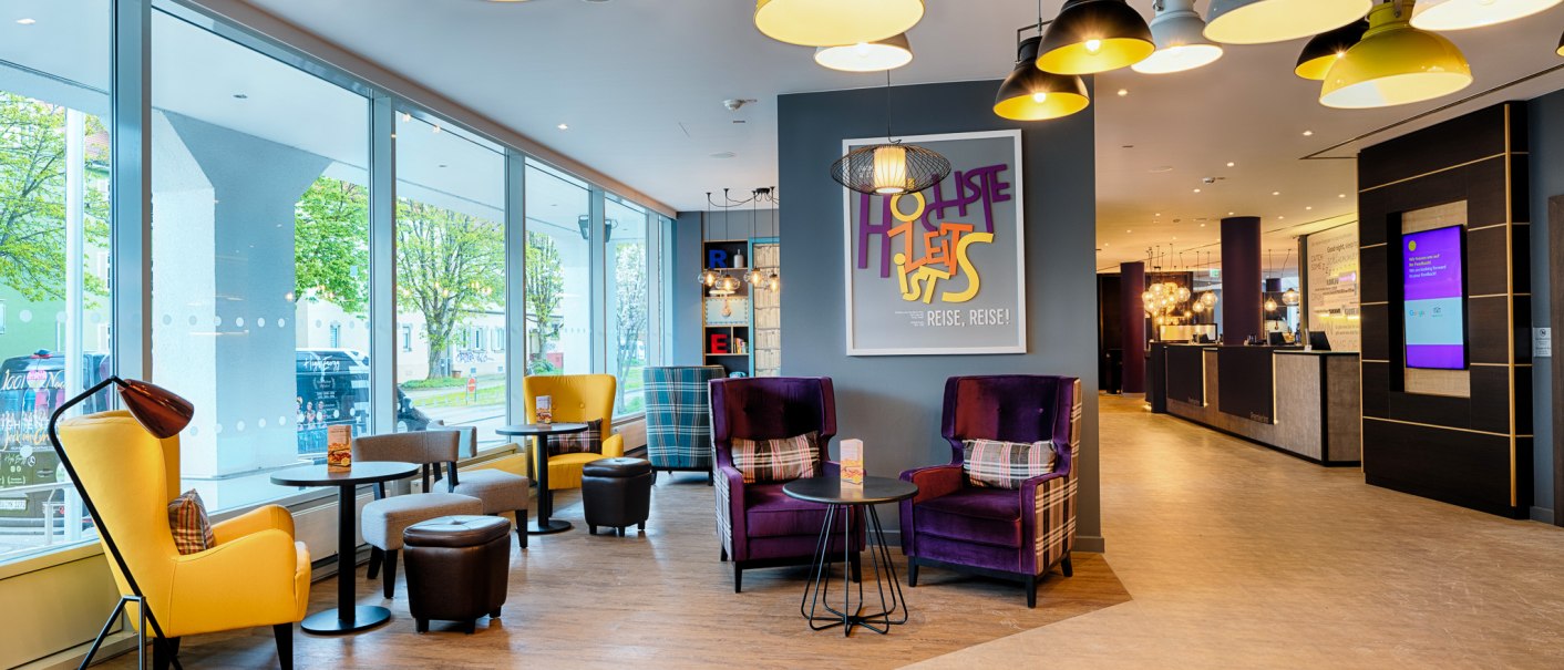 Moderne Lobby mit bunten Sesseln, großen Fenstern und stilvollen Lampen. Einladende Atmosphäre mit einem Empfangsbereich im Hintergrund., © Premier Inn Germany
