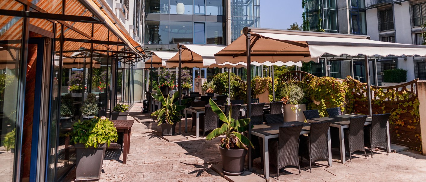 Sonniger Außenbereich eines Cafés mit Tischen, Stühlen und Pflanzen unter Markisen. Moderne Glasfassade im Hintergrund., © PLAZA Hotelgroup GmbH Sonniger Außenbereich eines Cafés mit Tischen, Stühlen und Pflanzen unter Markisen. Moderne Glasfassade im Hintergrund., © PLAZA Hotelgroup GmbH