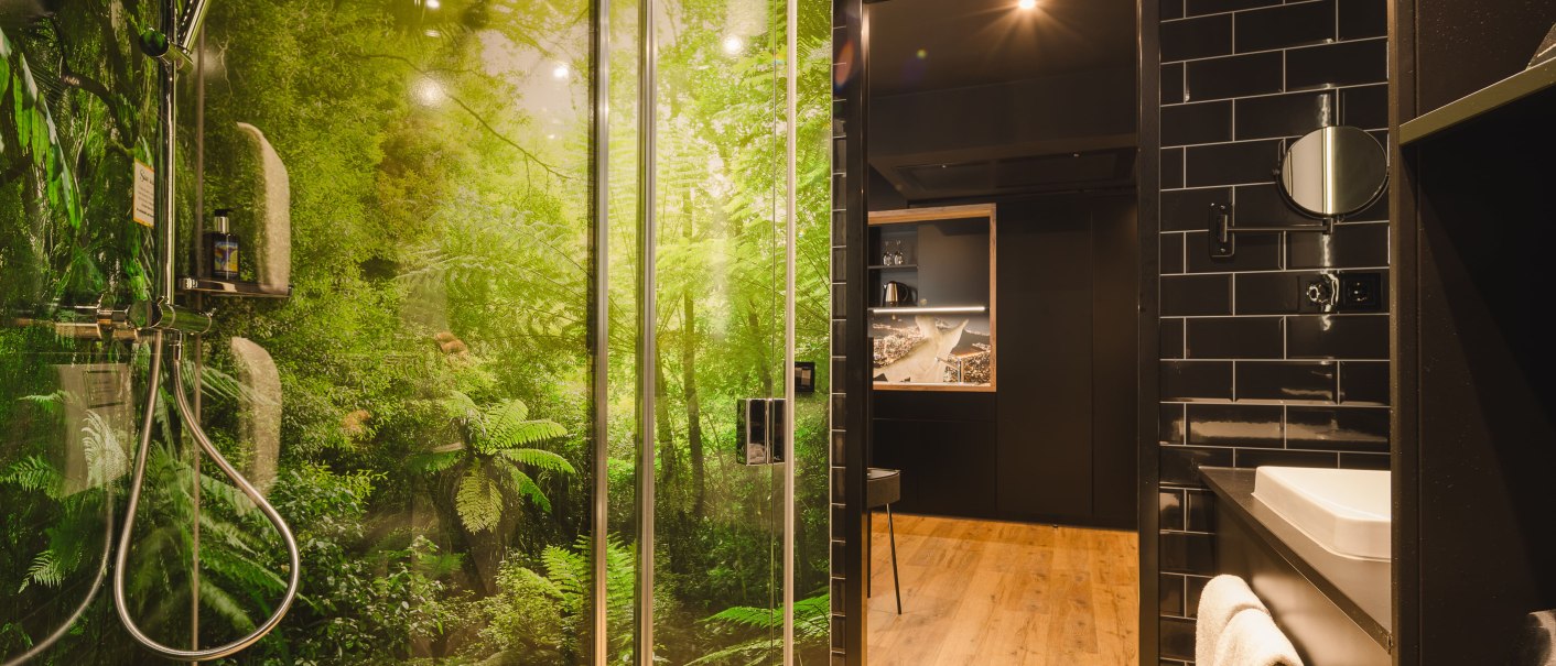 Modernes Badezimmer mit Dschungelmotiv in der Dusche, schwarzen Fliesen und Holzfußboden. Ein Waschbecken mit Handtuch ist sichtbar., © TOMAS Modernes Badezimmer mit Dschungelmotiv in der Dusche, schwarzen Fliesen und Holzfußboden. Ein Waschbecken mit Handtuch ist sichtbar., © TOMAS
