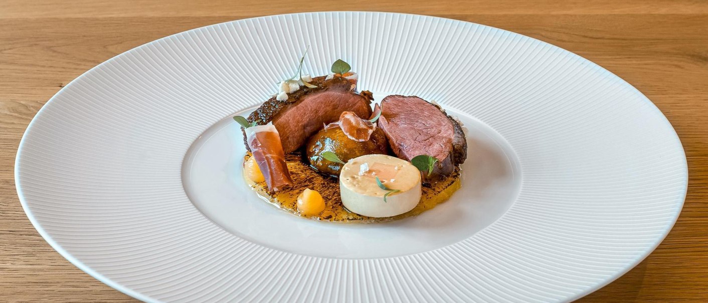 Gourmetgericht mit Entenbrust, dekorativ angerichtet auf einem weißen Teller. Beilagen und Soßen kunstvoll platziert, auf einem Holztisch serviert., © New Josch, Stuttgart