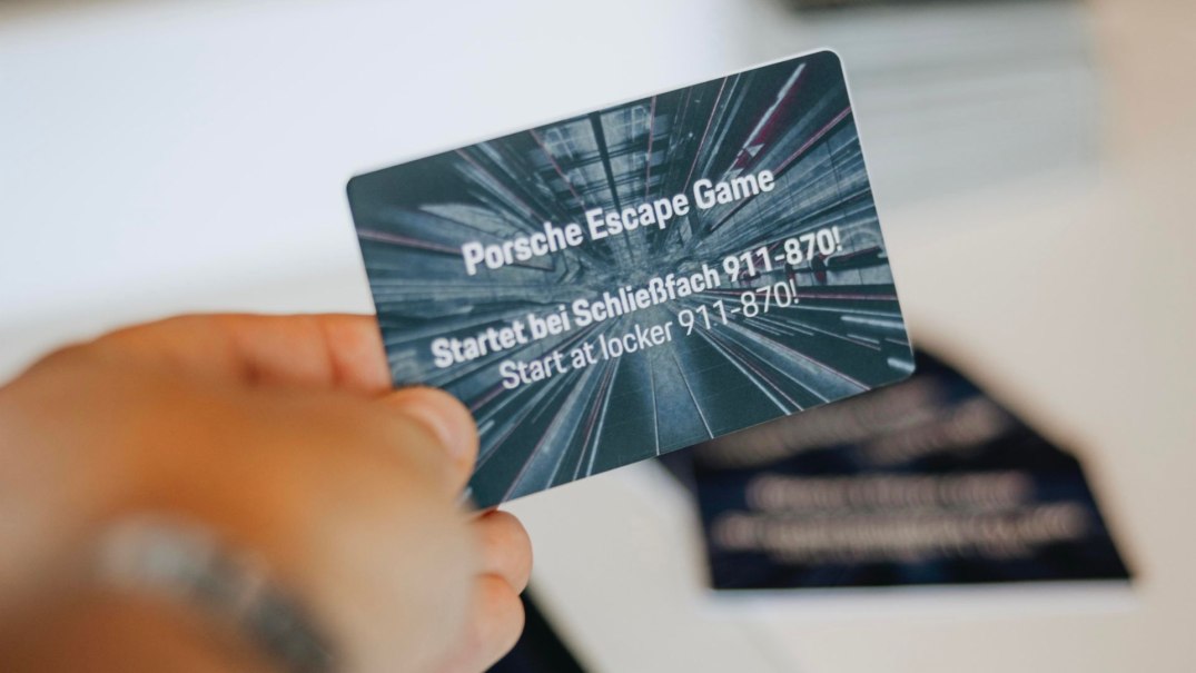 Eine Hand hält eine Karte mit der Aufschrift 'Porsche Escape Game. Startet bei Schließfach 911-870'. Hintergrund unscharf., © Porsche Museum