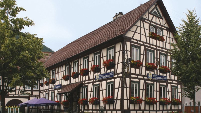 Fachwerkhaus mit roten Blumen an den Fenstern, Restaurant im Erdgeschoss, umgeben von Bäumen und einem sonnigen Himmel. Fachwerkhaus mit roten Blumen an den Fenstern, Restaurant im Erdgeschoss, umgeben von Bäumen und einem sonnigen Himmel.