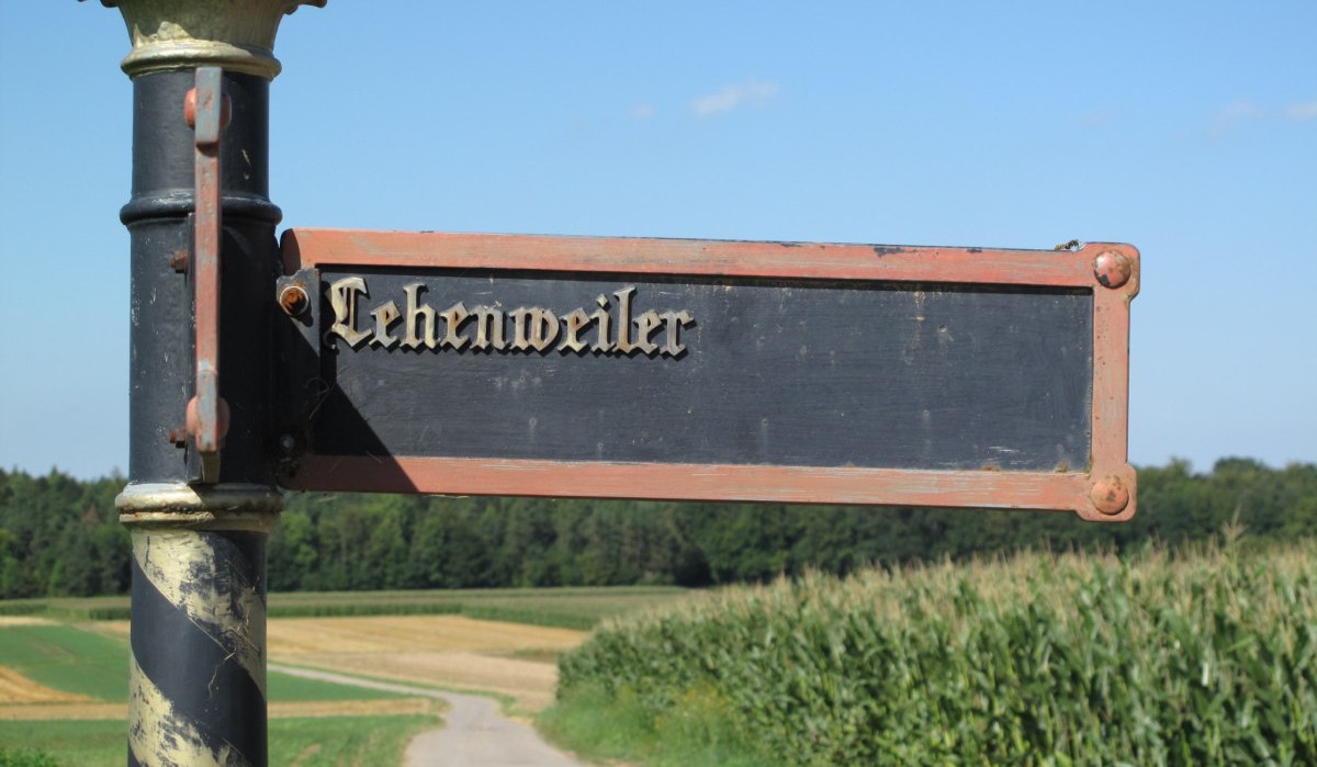 Ein altes Wegweiser-Schild mit der Aufschrift 'Lehenweiler' steht vor einem Feld und Wald unter klarem Himmel., &copy; Natur.Nah. Sch&ouml;nbuch & Heckeng&auml;u