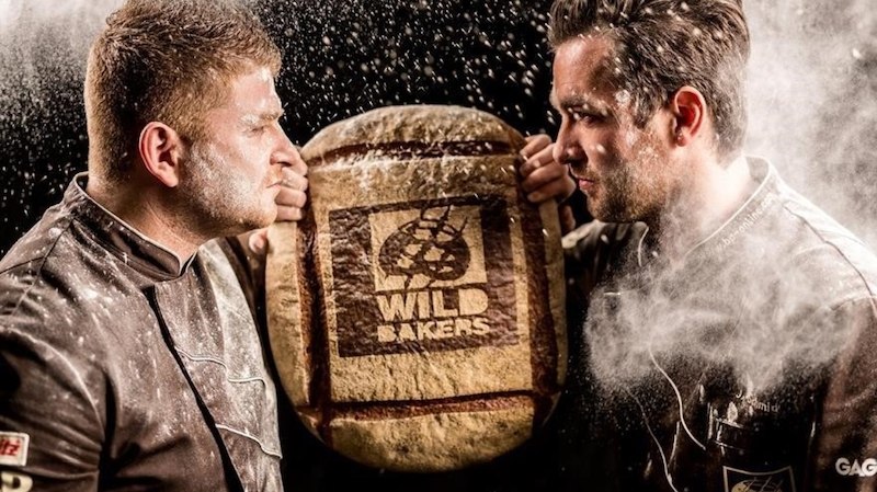 Zwei Bäcker stehen sich gegenüber, ein großes Brot mit dem Logo 'Wild Bakers' zwischen ihnen. Mehlstaub schwebt in der Luft., © Stadthalle Leonberg
