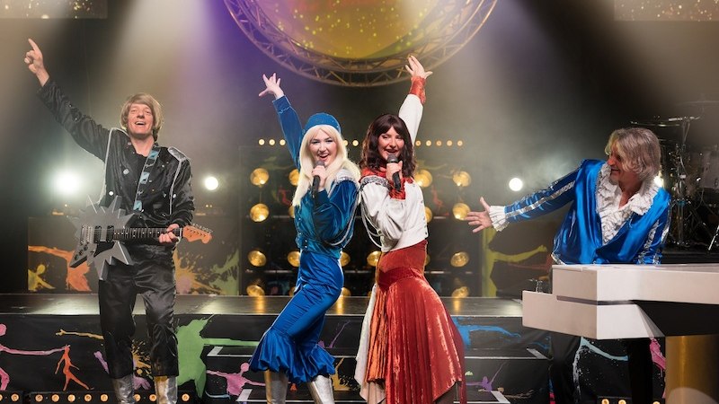 Vier K&uuml;nstler in bunten ABBA-Kost&uuml;men performen auf einer B&uuml;hne mit Lichteffekten. Im Hintergrund ist ein Schild mit 'GOLD' zu sehen., &copy; die Halle Kulturinitiative e.V.