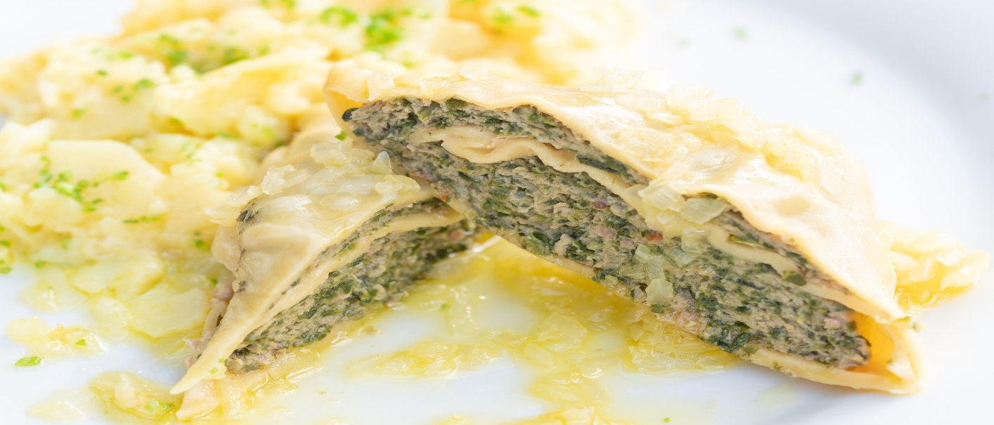 Nahaufnahme von Maultaschen mit einer Füllung aus Fleisch und Kräutern, serviert mit Kartoffelsalat auf einem weißen Teller., © Herr Kächele - Schwäbisches für Schleckige GmbH Nahaufnahme von Maultaschen mit einer Füllung aus Fleisch und Kräutern, serviert mit Kartoffelsalat auf einem weißen Teller., © Herr Kächele - Schwäbisches für Schleckige GmbH