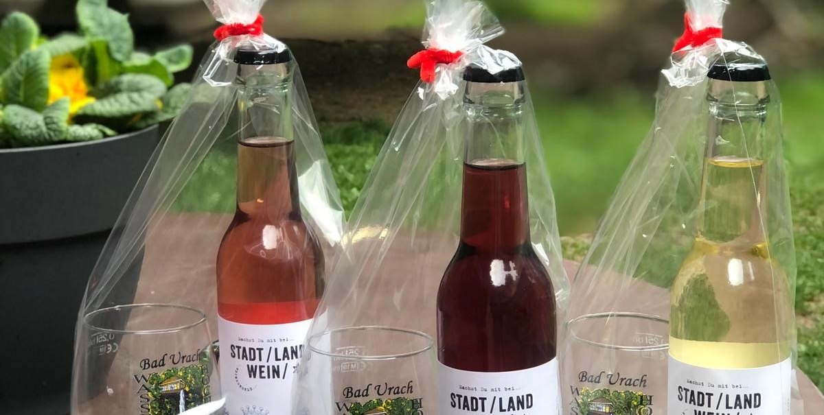 Vier Flaschen Stadt/Land Wein in Geschenkverpackung stehen auf einem runden Tisch im Freien. Im Hintergrund ist grüne Vegetation zu sehen., © n/a