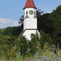 Ein Kirchturm mit rotem Dach und Uhr ragt über eine bunte Blumenwiese, umgeben von grünen Bäumen, in den blauen Himmel. Ein Kirchturm mit rotem Dach und Uhr ragt über eine bunte Blumenwiese, umgeben von grünen Bäumen, in den blauen Himmel.