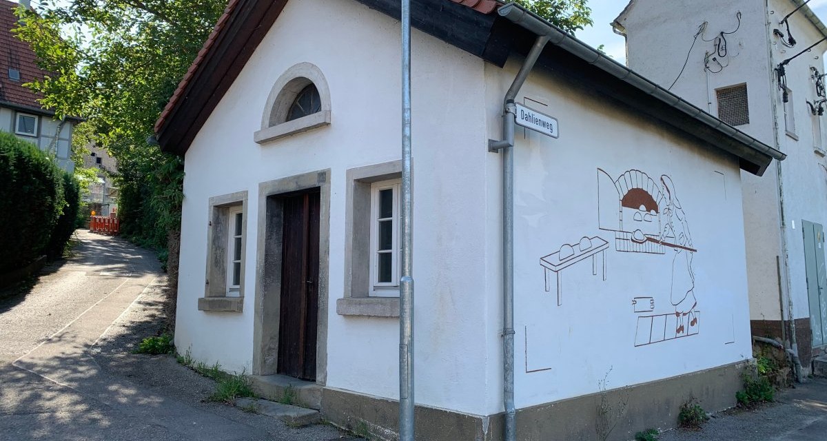 Kleines wei&szlig;es Geb&auml;ude mit rotem Dach, Wandgem&auml;lde eines B&auml;ckers, Stra&szlig;enschild 'Dahlienweg', B&auml;ume im Hintergrund., &copy; Land der 1000 H&uuml;gel - Kraichgau-Stromberg