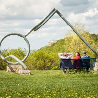 Drei Personen sitzen auf einer Bank vor einer großen Brillenskulptur in einer grünen Landschaft mit Hügeln und Wolken am Himmel., © hochgehberge
