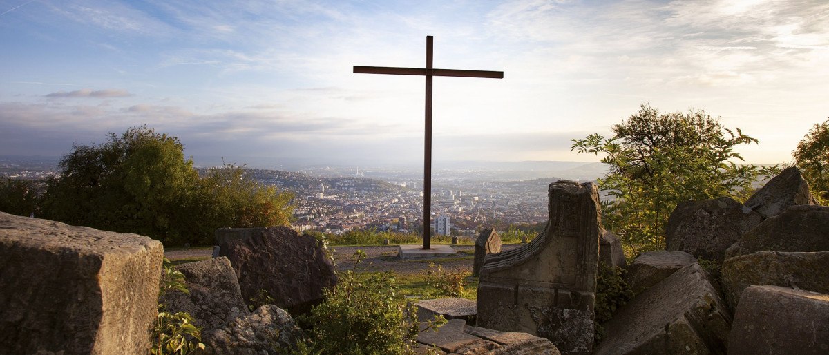 Holzkreuz auf dem Birkenkopf in Stuttgart, umgeben von Trümmern, mit Blick auf die Stadt im Hintergrund bei Sonnenuntergang., © Stuttgart-Marketing GmbH Holzkreuz auf dem Birkenkopf in Stuttgart, umgeben von Trümmern, mit Blick auf die Stadt im Hintergrund bei Sonnenuntergang., © Stuttgart-Marketing GmbH