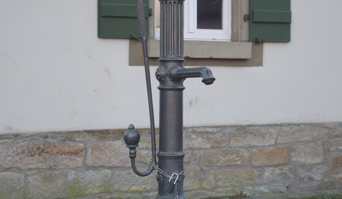 Ein alter Pumpbrunnen aus Metall steht vor einem Gebäude mit grünen Fensterläden und einem steinernen Sockel., © Land der 1000 Hügel - Kraichgau-Stromberg Ein alter Pumpbrunnen aus Metall steht vor einem Gebäude mit grünen Fensterläden und einem steinernen Sockel., © Land der 1000 Hügel - Kraichgau-Stromberg