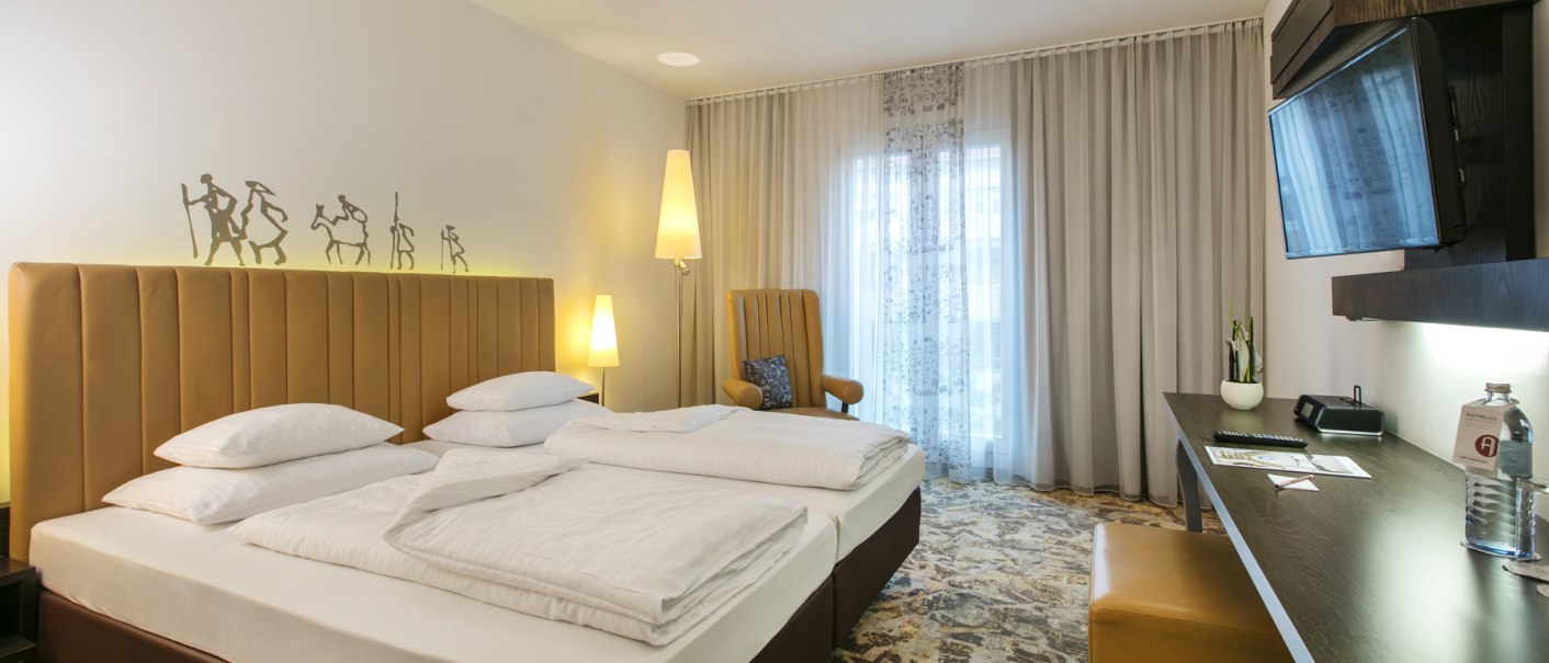 Modernes Hotelzimmer mit Doppelbett, Schreibtisch, Fernseher und Sessel. Dezente Wanddekoration und große Fenster mit Vorhängen., © ARCOTEL Hotels