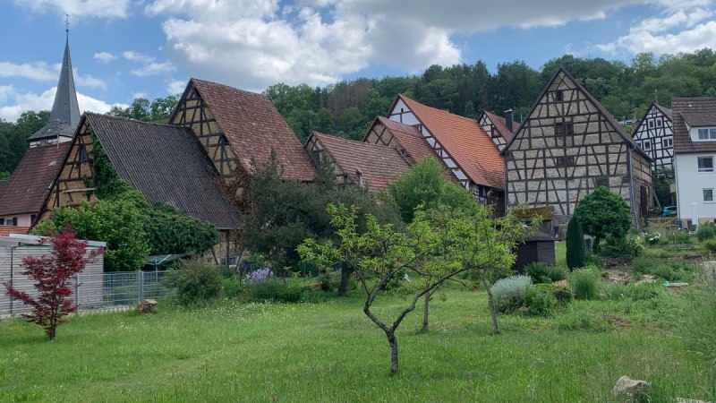 Fachwerkhäuser mit roten Dächern und ein Kirchturm in einem Dorf, umgeben von grüner Landschaft und Bäumen., © Land der 1000 Hügel - Kraichgau-Stromberg Fachwerkhäuser mit roten Dächern und ein Kirchturm in einem Dorf, umgeben von grüner Landschaft und Bäumen., © Land der 1000 Hügel - Kraichgau-Stromberg