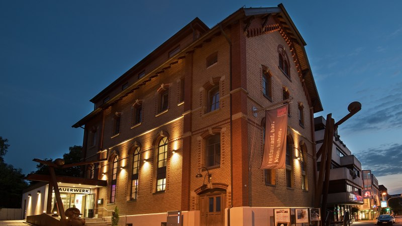 Das Mauerwerk in Herrenberg bei Dämmerung, ein beleuchtetes Backsteingebäude mit großen Fenstern und einem Banner. Der Himmel ist dunkelblau., © Stadt Herrenberg Das Mauerwerk in Herrenberg bei Dämmerung, ein beleuchtetes Backsteingebäude mit großen Fenstern und einem Banner. Der Himmel ist dunkelblau., © Stadt Herrenberg