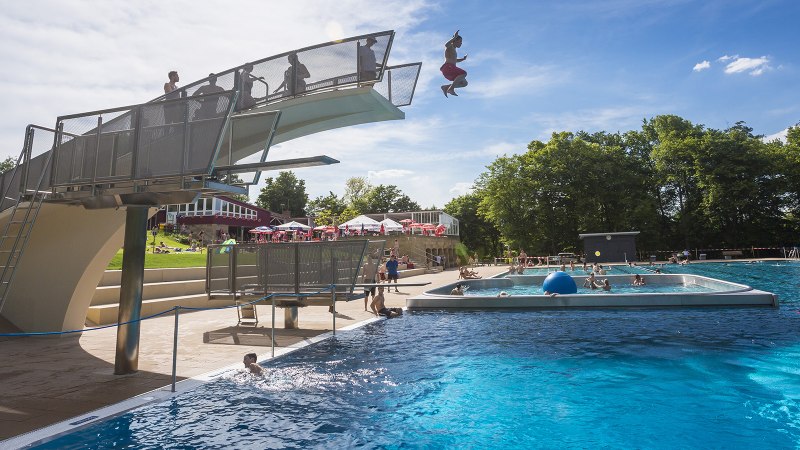 Ein Freibad mit Sprungturm, von dem ein Mensch ins Wasser springt. Viele Menschen schwimmen im großen Becken. Im Hintergrund sind Bäume und Sonnenschirme., © Stuttgarter Bäder