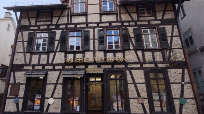 Fachwerkhaus mit der Aufschrift 'Galerie Wiedmann'. Das Geb&auml;ude hat mehrere Fenster mit gr&uuml;nen Fensterl&auml;den und eine Laterne an der Seite., &copy; Galerie Wiedmann