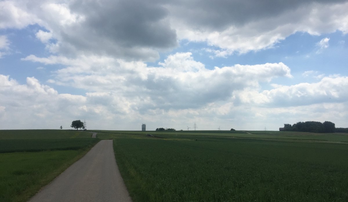 Landschaft mit schmalem Weg, grünen Feldern und einem Wasserturm in der Ferne unter einem bewölkten Himmel., © www.pro-cycl.de Landschaft mit schmalem Weg, grünen Feldern und einem Wasserturm in der Ferne unter einem bewölkten Himmel., © www.pro-cycl.de