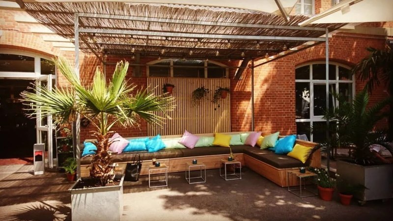 Terrasse mit bunten Kissen auf einer Eckbank, Palmen in Töpfen und einem Holzdach. Ziegelwand im Hintergrund., © Blauer Engel GmbH Terrasse mit bunten Kissen auf einer Eckbank, Palmen in Töpfen und einem Holzdach. Ziegelwand im Hintergrund., © Blauer Engel GmbH