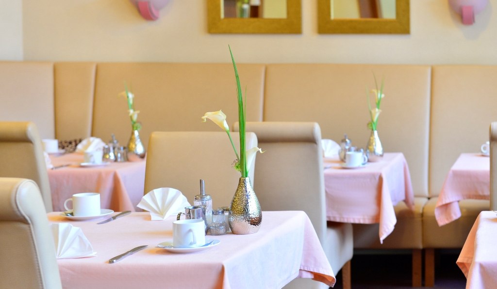 Eleganter Frühstücksraum mit rosa Tischdecken, weißen Tassen und Blumenvasen. Beige Sitzbänke und Spiegel an der Wand., © Hotel Fuchsen Eleganter Frühstücksraum mit rosa Tischdecken, weißen Tassen und Blumenvasen. Beige Sitzbänke und Spiegel an der Wand., © Hotel Fuchsen
