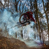 Ein Mountainbiker in rotem Karohemd springt durch Rauch im Wald. Die Szene ist dynamisch und zeigt herbstliche Bäume im Hintergrund., © Natur.Nah. Schönbuch & Heckengäu