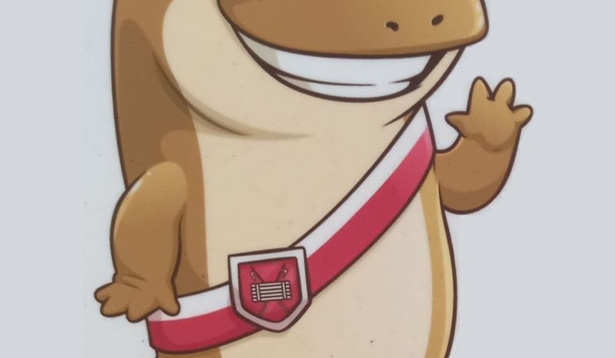 Ein lächelndes, cartoonartiges Tier mit großen Augen, rotem Band und Wappen, das freundlich winkt.