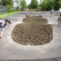 Fahrradfahrer auf einem Pumptrack in Sindelfingen, umgeben von Zuschauern und Bäumen. Die Strecke ist asphaltiert und wellig., © Fotoknobi