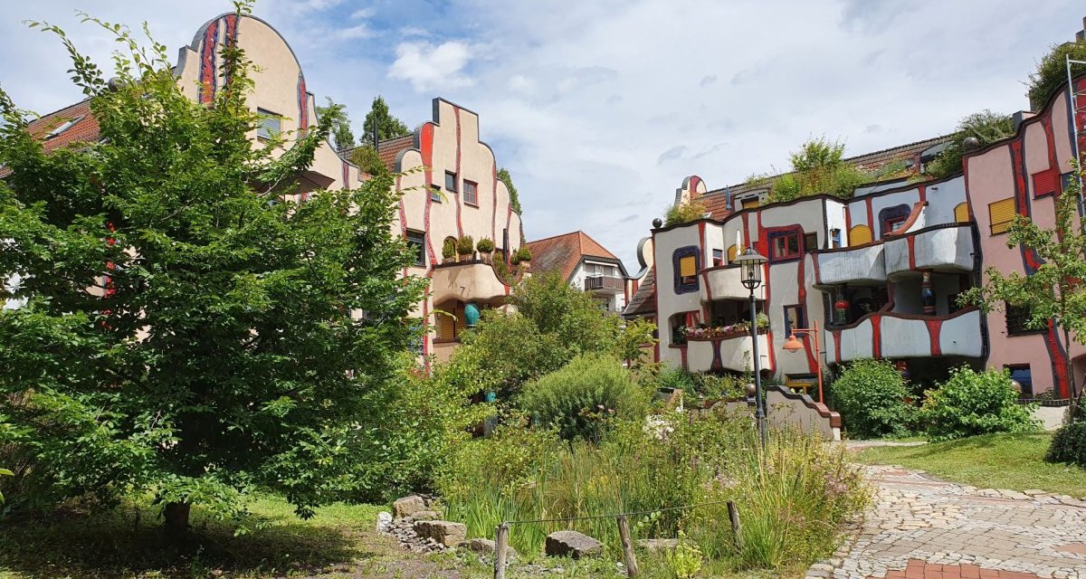 Buntes Gebäude im Hundertwasser-Stil mit geschwungenen Linien, Balkonen und viel Grün im Vordergrund. Ein gepflasterter Weg führt durch den Garten. Buntes Gebäude im Hundertwasser-Stil mit geschwungenen Linien, Balkonen und viel Grün im Vordergrund. Ein gepflasterter Weg führt durch den Garten.