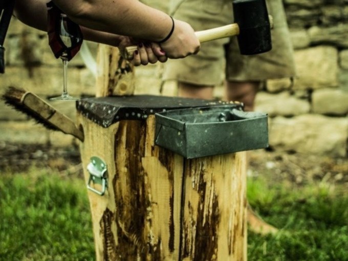 Eine Person hält einen Hammer und ein Weinglas neben einem Holzblock im Freien. Im Hintergrund ist eine Steinmauer zu sehen., © Cool-Tours StattReisen Eine Person hält einen Hammer und ein Weinglas neben einem Holzblock im Freien. Im Hintergrund ist eine Steinmauer zu sehen., © Cool-Tours StattReisen