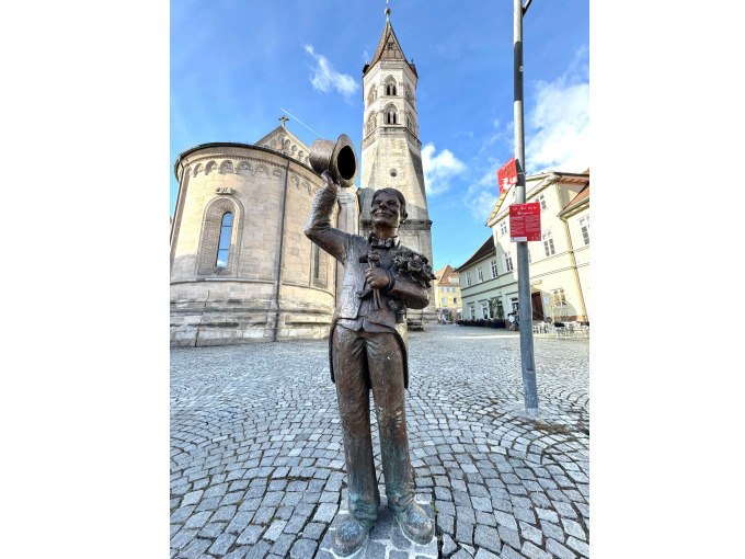 Bronzestatue eines Mannes mit Zylinder und Blumenstrauß vor einer Kirche mit Turm. Der gepflasterte Platz und der blaue Himmel runden die Szene ab., © Touristik und Marketing GmbH Bronzestatue eines Mannes mit Zylinder und Blumenstrauß vor einer Kirche mit Turm. Der gepflasterte Platz und der blaue Himmel runden die Szene ab., © Touristik und Marketing GmbH