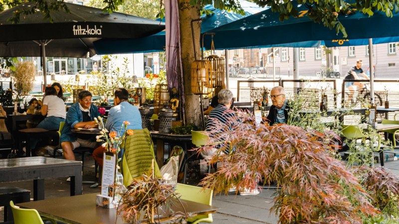 Ein belebtes Café im Freien mit Menschen, die an Tischen unter Sonnenschirmen sitzen. Pflanzen und Dekorationen schaffen eine gemütliche Atmosphäre., © SMG Stuttgart Marketing GmbH - Sarah Schmid