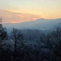 Sonnenuntergang über nebliger Landschaft, Bäume im Vordergrund, Hügel im Hintergrund. Der Himmel ist in warmen Farben gefärbt.