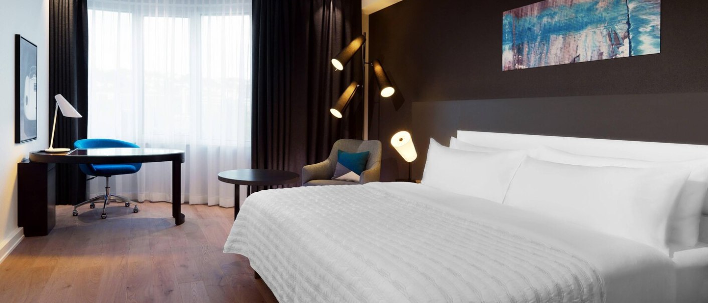 Modernes Hotelzimmer mit großem Bett, Schreibtisch, blauem Stuhl und Sessel. Helle Vorhänge und stilvolle Beleuchtung., © Hotel Le Méridien