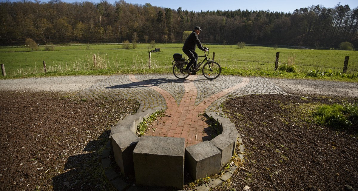 Ein Radfahrer fährt auf einem gepflasterten Weg durch eine grüne Landschaft mit Wiesen und Wäldern im Hintergrund., © VRS/ Stoppel Ein Radfahrer fährt auf einem gepflasterten Weg durch eine grüne Landschaft mit Wiesen und Wäldern im Hintergrund., © VRS/ Stoppel