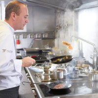 Ein Koch in weißer Uniform brät Fleisch in einer Pfanne in einer professionellen Küche. Im Hintergrund ist ein weiterer Koch zu sehen., © SMG, Jean-Claude Winkler