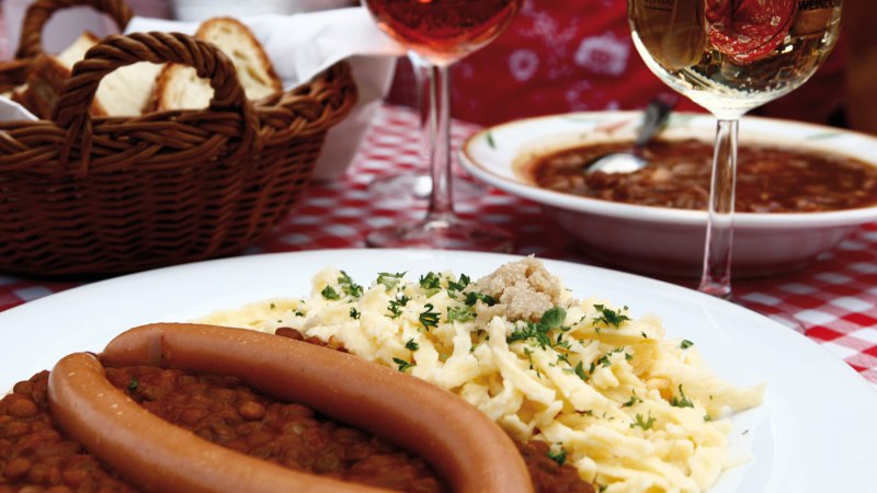 Ein Teller mit Linsen, Spätzle und Würstchen auf einem rot-weiß karierten Tischtuch. Im Hintergrund ein Brotkorb und ein Glas Wein., © Stuttgart-Marketing GmbH Ein Teller mit Linsen, Spätzle und Würstchen auf einem rot-weiß karierten Tischtuch. Im Hintergrund ein Brotkorb und ein Glas Wein., © Stuttgart-Marketing GmbH