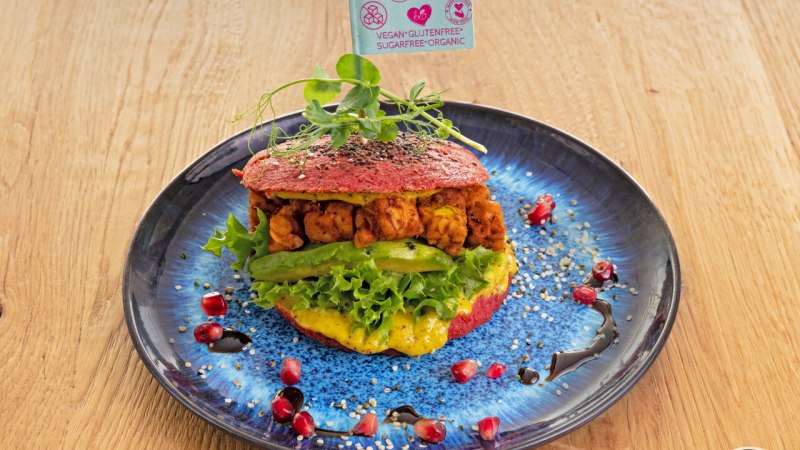 Ein veganer Burger mit Salat und Avocado auf einem blauen Teller, dekoriert mit Granatapfelkernen und Sesam. Ein Fähnchen weist auf vegane, glutenfreie Zutaten hin., © cEnergeticLife Ein veganer Burger mit Salat und Avocado auf einem blauen Teller, dekoriert mit Granatapfelkernen und Sesam. Ein Fähnchen weist auf vegane, glutenfreie Zutaten hin., © cEnergeticLife