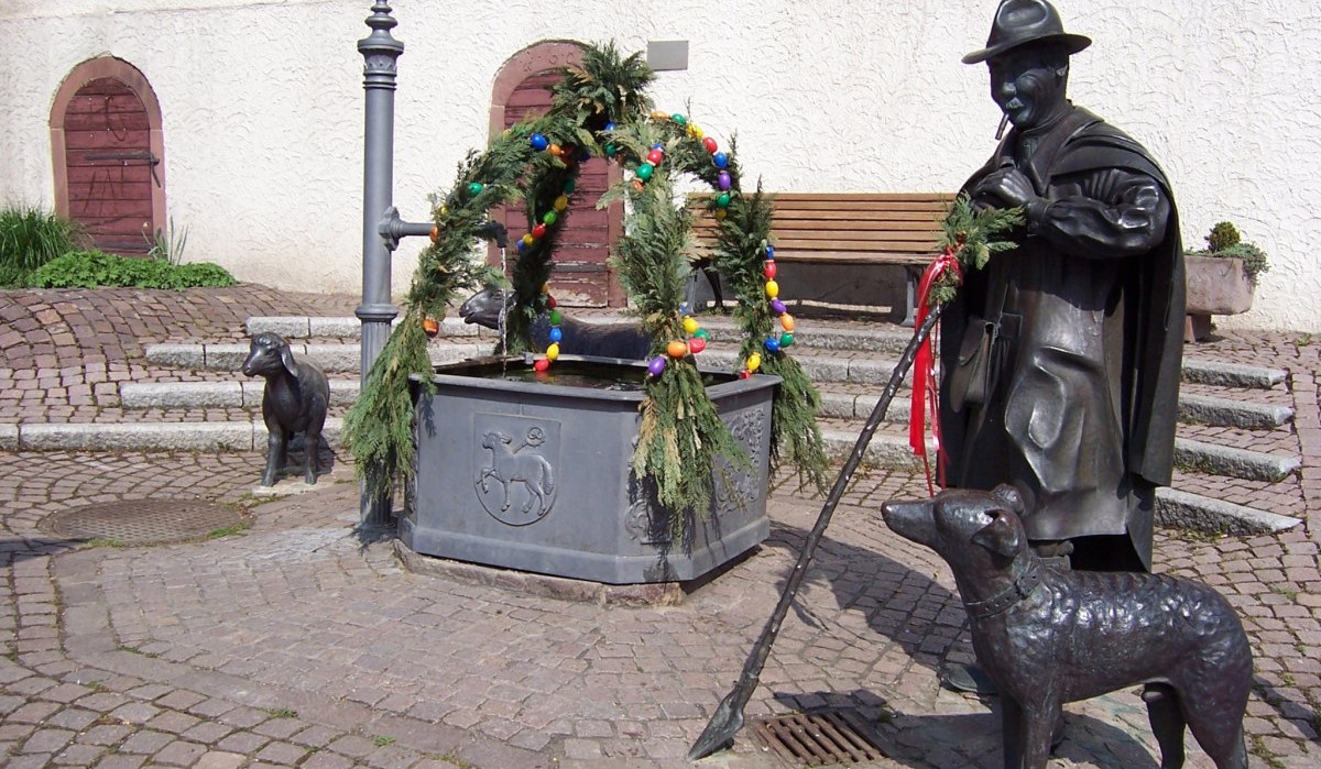 Der Osterbrunnen in Schafhausen ist mit bunten Eiern und gr&uuml;nen Zweigen geschm&uuml;ckt. Bronzefiguren eines Hirten, eines Hundes und eines Schafes umgeben den Brunnen., &copy; Natur.Nah. Sch&ouml;nbuch & Heckeng&auml;u