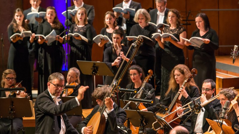 Ein Chor und Orchester in formeller Kleidung führen ein klassisches Musikstück auf. Der Dirigent leitet die Musiker, die konzentriert spielen und singen., © Holger Schneider
